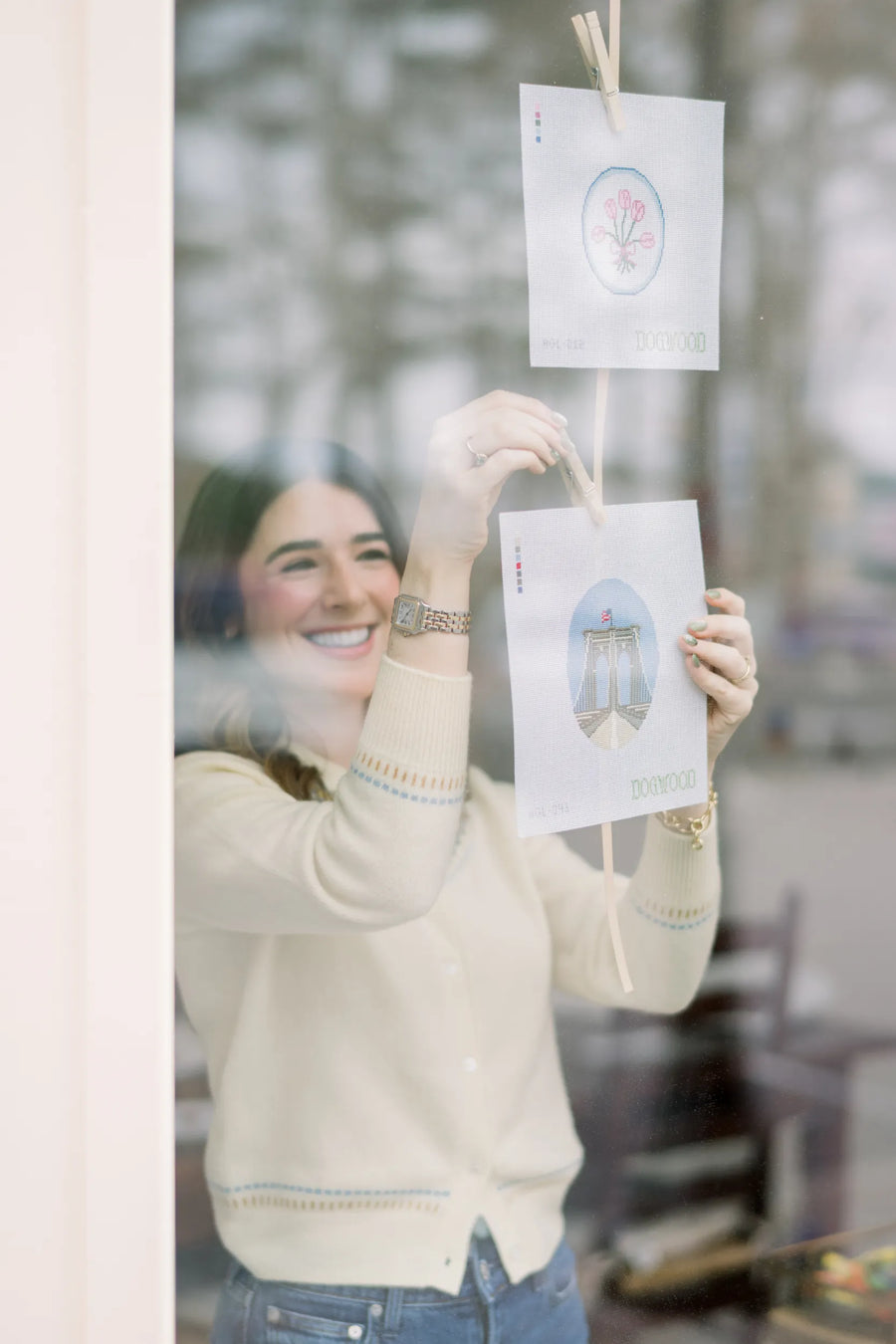 Abbie Lilly hanging Dogwood Needlepoint tulip and Brooklyn Bridge canvases in a window with clothespins