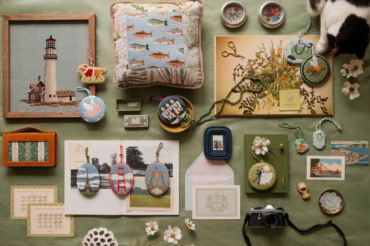 Flat lay of Dogwood Needlepoint canvases, ornaments, and stitching tools styled with dried flowers and a cat.