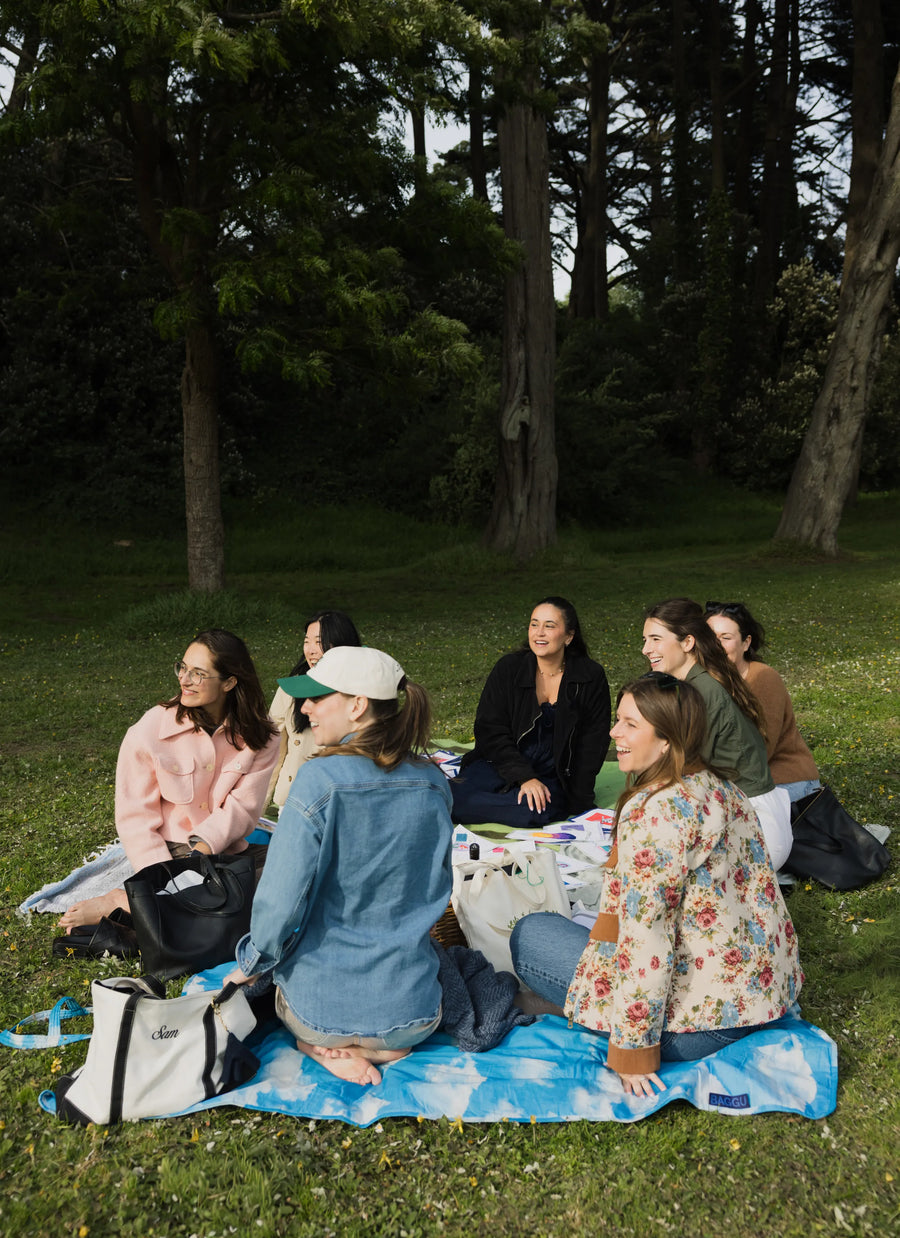 A group of women sitting on a blue and white picnic blanket on a grassy lawn in a park, surrounded by trees.