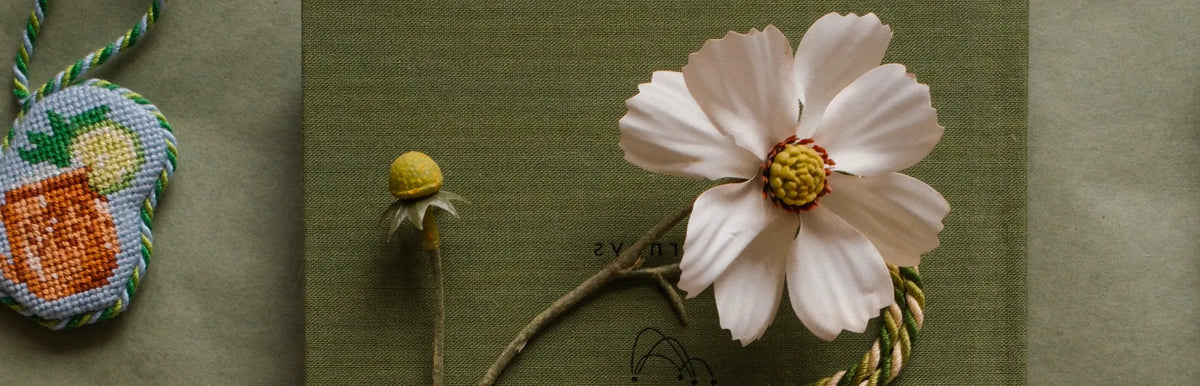 Dogwood Needlepoint stockists page header featuring a pressed cosmos flower and citrus-print needlepoint ornament on olive green fabric and paper.